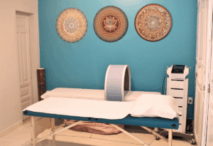 Sala de terapias integrativas Sadhu com maca de atendimento, aparelho terapêutico circular sobre a maca e mandalas decorativas na parede azul.