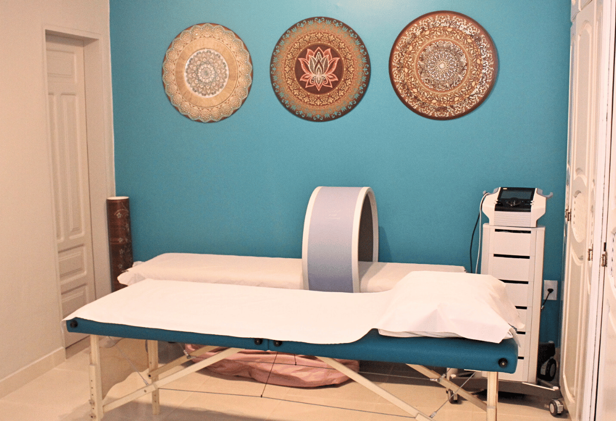 Sala de terapias integrativas Sadhu com maca de atendimento, aparelho terapêutico circular sobre a maca e mandalas decorativas na parede azul.